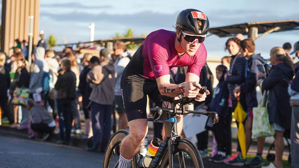 A cyclist with a Breathy nasal strip in a pink jersey and black shorts rides a road bike on an asphalt road during a race, with a crowd of spectators lining the side under a cloudy sky.